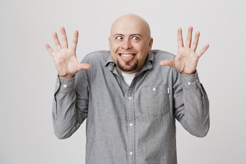Naklejka premium Indoor shot of bald bearded man making grimace with slanting eyes, posing against gray background with opened palms, sticking out his tongue. Positive face expression and emotions