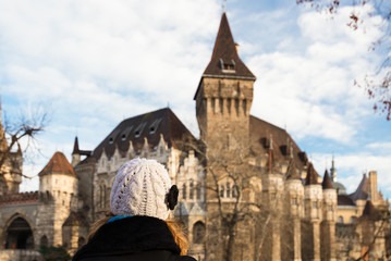 Fototapeta premium Young woman is looking at the Vajdahunyad Castle (Hungarian Agricultural Museum) which i one of the romantic castles in Budapest, Hungary, located in the City Park by the boating lake / skating rink.
