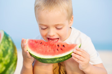 Handsome boy eating slice of watermelon on the beach. Summertime. The concept of a happy childhood.
