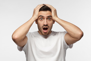Closeup portrait of emotional man isolated on gray background with strong expression of fear, round eyes, open mouth and hands pressed to top of head, showing astonishment and shock - obrazy, fototapety, plakaty