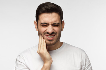 Fototapeta premium Closeup of young man isolated on gray background touching his face and closing eyes with expression of horrible suffer from health problem and aching tooth, showing dissatisfaction