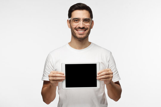Studio picture of positive man isolated on grey background standing in casual clothes holding tablet and showing it blank screen with happy smile as if advising product or service