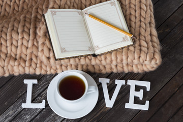 on a dark wooden floor a brown warm plaid, a notepad, a pencil, a cup of tea and the word love from wooden letters