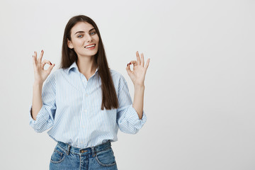 Portrait of satisfied female teacher, showing okay or excellent gesture with both hands, smiling at camera while standing over gray background. Student shows she passed her tests with body language.