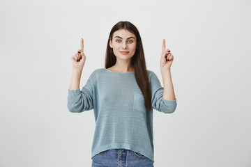 Pretty smiling joyfully female with dark long hair, pointing with fingers up, showing copy space for advertising content. Good-looking beautiful woman advertising something, isolated on studio wall.