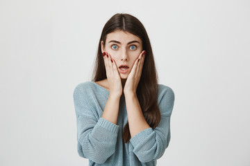 Shocked stunned emotional woman with long straight hair keeps hands on cheeks, is troubled and frustrated, isolated against gray background. European female can`t believe in shocking news