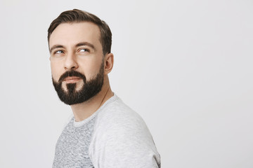 Obraz premium Side view of attractive thoughtful bearded man, turned over to look a something suspiciously, standing against gray background. Guy forgot glasses at home and can not what time is it