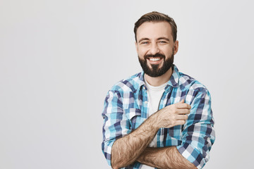 Portrait of adult bearded man with shiny smile and crossed hands, looking at camera, over gray...