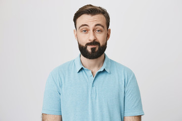 Portrait of attractive adult with beard and moustache expressing that he is not impressed, lifting his eyebrows and smiling, over gray background. Guy knows he hears lie but makes look he believes.