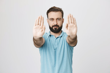 Studio shot of attractive young man with beard and stylish haircut stretching his palms at camera with calm expression, over gray background. Guy makes morning exercises indoor.