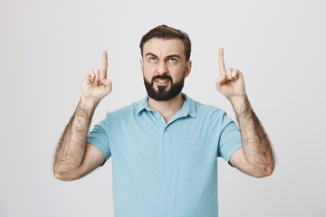 Indoor portrait of angry european bearded male pointing up with index fingers and gloomy smile, over gray background. Man hate his annoying neighbors who always yell at midnight