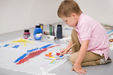 Cute kid boy sitting on the floor and drawing with colorful paints.Child making flag craft. Education, creativity and school activities.