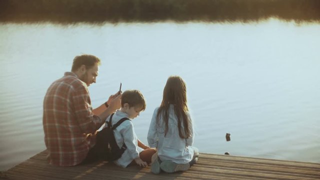 Father and two kids sit together on a lake pier. European man with children photographs water birds with smartphone. 4K.
