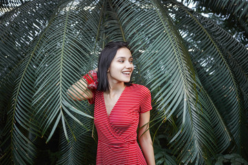 Horizontal view of positive young Caucasian brunette female in fashionable red dress with white stripes posing against lush of green tropical plants, having joyful happy expression on her cute face