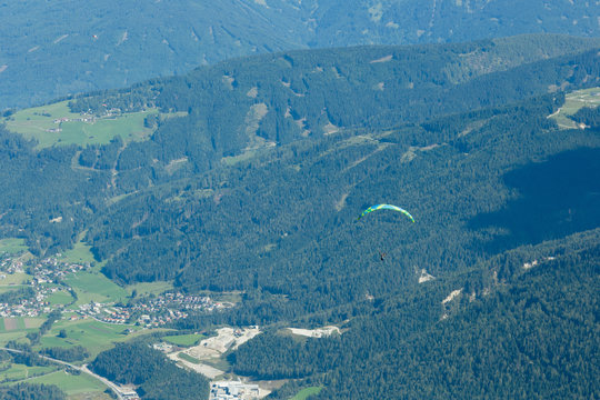A paraglider looking down on a village soaring in the Austrian Alps. The paraglider is shown above a forest high above ground level.
