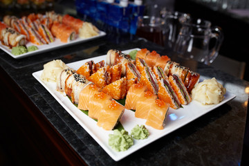 Eating sushi rolls. Japanese food restaurant, sushi maki gunkan roll plate or platter set. Closeup of hand with chopsticks taking roll. Ginger, soy, wasabi. Sushi at black rustic wood background.