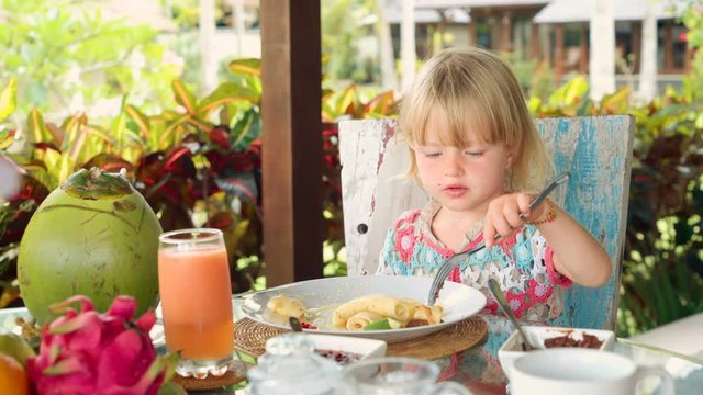 Cute little caucasian girl eating thin pancakes with fork for breakfast