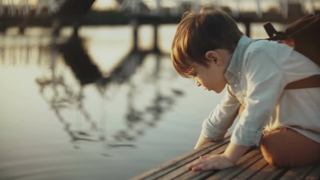 Cute European boy plays with lake water on a pier. Little male child with backpack exploring summer nature. 4K close-up.