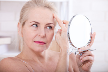 Middle aged woman looking at wrinkles in mirror. Plastic surgery and collagen injections. Makeup....