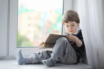 The little boy is reading a book. The child sits at the window a