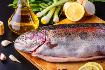 Fresh gutted trout on a wooden cutting board surrounded by ingredients for baking: olive oil, lemon, spring onions, garlic and spices. Top view