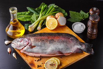 Fresh gutted trout on a wooden cutting board surrounded by ingredients for baking: olive oil, lemon, spring onions, garlic and spices. Top view