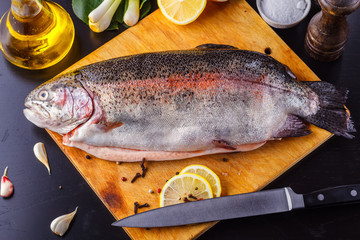 Fresh gutted trout on a wooden cutting board surrounded by ingredients for baking: olive oil, lemon, spring onions, garlic and spices. Top view