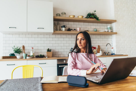 Portrait Of Young Woman Sitting At The Table In Kitchen Working On Laptop.