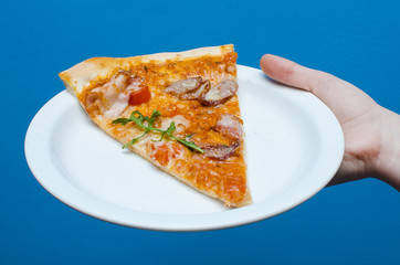 In the hand a plate with delicious pizza with meat and greens on a blue background
