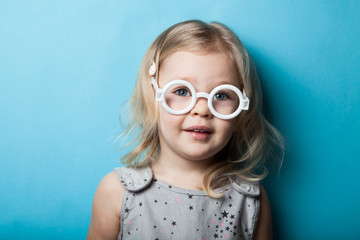 A small European girl in white toy glasses is isolated on a blue background. Portrait.