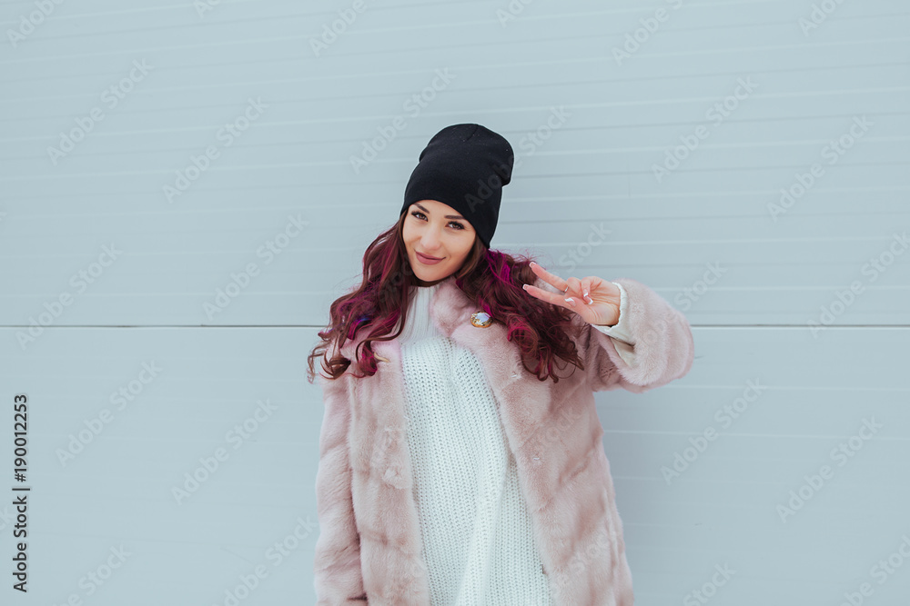 Wall mural Portrait of beauty fashion smiling woman with colored ombre hairstyle, making peace by fingers in mink coat on grey background. Outdoor