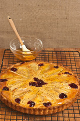 Front view of a freshly baked, homemade, pear cranberry tart with almond glaze cooling on a wire  cooling rack on a wood serving board with a glass bowl of glaze and a wood application brush
