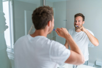 Man getting ready in the morning doing hygiene routine brushing his teeth looking in mirror of home bathroom using toothbrush in for clean dental oral care.