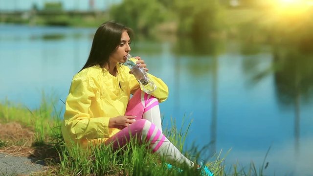 Girl sits on grass near lake and drinks water. Attractive woman in sportswear drinks clean water from bottle. Girl opens bottle of water and drinks soda water, beautiful sun with flicker at background