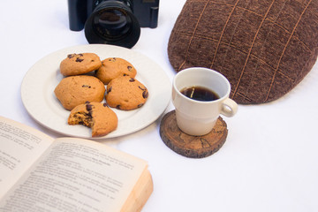 Table with chocolate chips cookies, book, cup of coofee, camera and hat