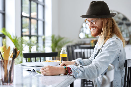 Cheerful bearded male guy in stylish denim jacket, hat and eyewear reads magazines while sits at kitchen table, going to have lunch, reads very funny article. Hipster guy enjoys free time, reads