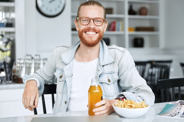 Indoor shot of bearded ginger male in eyewear enjoys dinner time at cozy restaurant, eats fast food and juice, has happy expression. Hipster guy has spare time after work, spends time at cafe