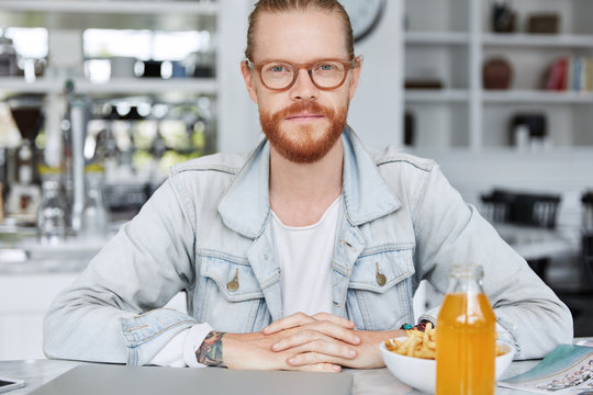 Confident redhaired male with beard and mustche spends free time at cafeteria, eats junk food, looks directrly into camera as sits at table with food, drink and modern portable laptop computer