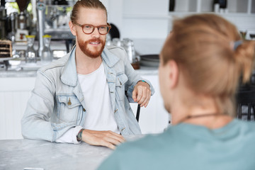Talented male designer with ginger beard and mustache, dressed in denim jacket, wears spectalces, has meeting with colleague, discuss future project together, share their experience or ideas.