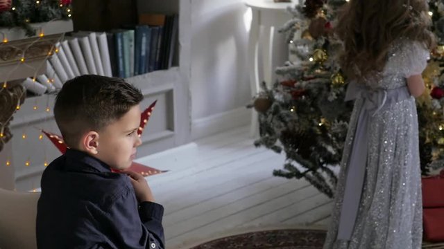 boy and girl in a christmas interior. the boy is sitting on the armchair, the girl is standing near the Christmas tree.