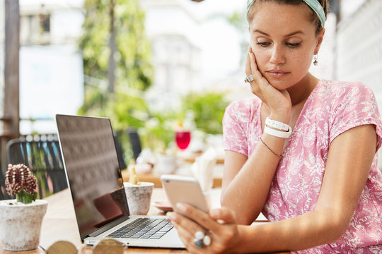 Serious freckled female model rests in outdoor cafe, waits her order, reads attentively local news on smart phone, laptop computer in front for distance work. Woman looks weather forecast on cellular
