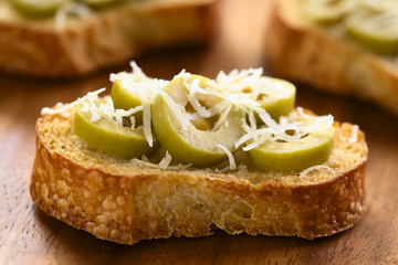 Frische Crostini oder geröstete Brotscheiben mit grünen Oliven und geriebenen Käse, fotografiert mit natürlichem Licht (Selektiver Fokus, Fokus auf die Vorderkante der vorderen Olivenscheiben)