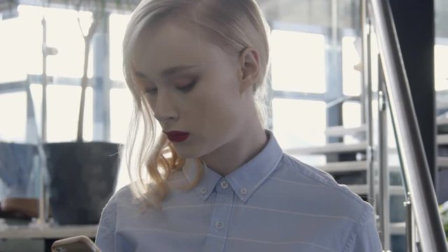 Portrait of beautiful female blonde who is seriously reading the phone. Young woman in a stylish shirt in public place holds smartphone in her hands and reads an SMS with a considerate expression.