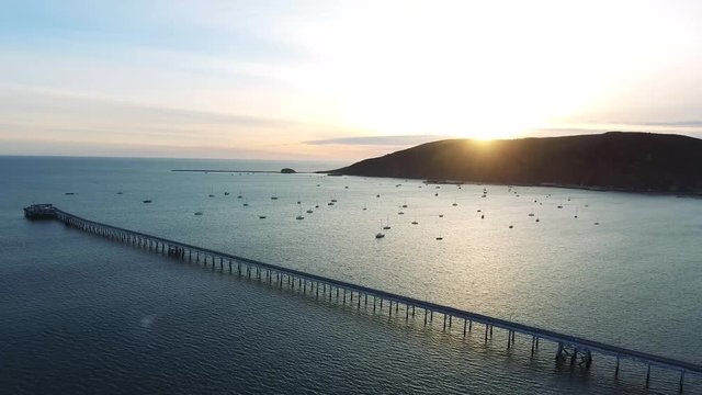 Aerial Drone Flyover - Panning left in a Pacific ocean bay at sunset. In the scene is a pier, sailboats, mountains and sky.