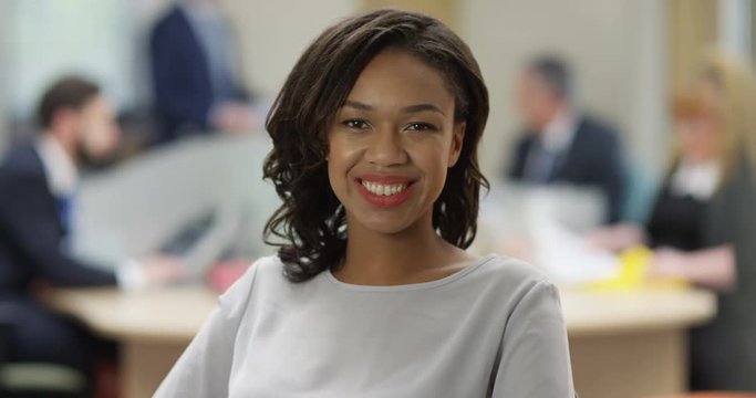 4K Portrait attractive mixed race businesswoman with a friendly smile & the rest of her team working in background. Slow motion.