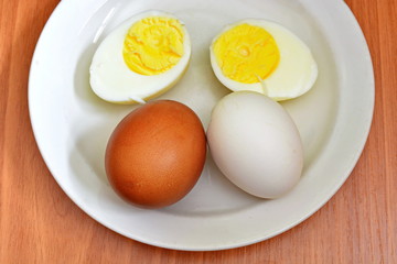 Half hard boiled chicken egg and whole red and white egg in eggshell on a white saucer and wooden table
