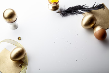 Fototapeta premium Easter holiday composition with eggs painted golden, yolk in egg stand, black feather and golden ribbon. White background, copy space