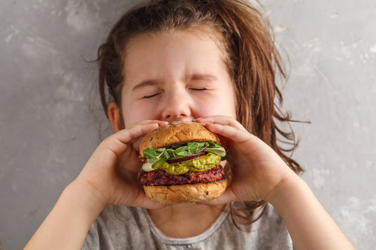 Beautiful happy hungry baby girl eating vegan burger.  Vegan beet chickpea burgers with vegetables, guacamole and rye buns. Healthy child vegan food concept.