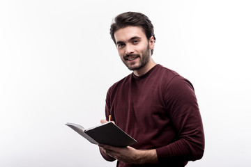 Important remarks. Charming young man taking notes in his daily planner and smiling at the camera while posing against a white background