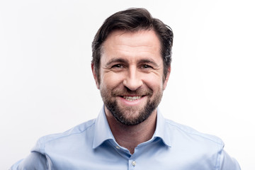 Appealing smile. The portrait of a handsome dark-haired middle-aged man in a blue shirt smiling at the camera while posing against a white background